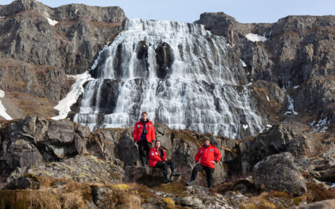 3 Iceland travelers in red jackets pose in front of the Dynjandi waterfall with cascading water among rocky cliffs, in the sun.