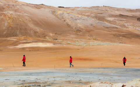 3 Iceland travelers in red jackets walk among ochre hills & gray boiling thermal springs under a hazy sky.