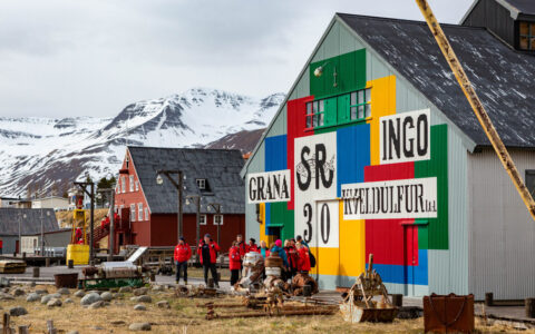 Small group of Iceland cruise guests in red parkas walk among brightly-painted metal buildings in a seaside village by snowy peaks.