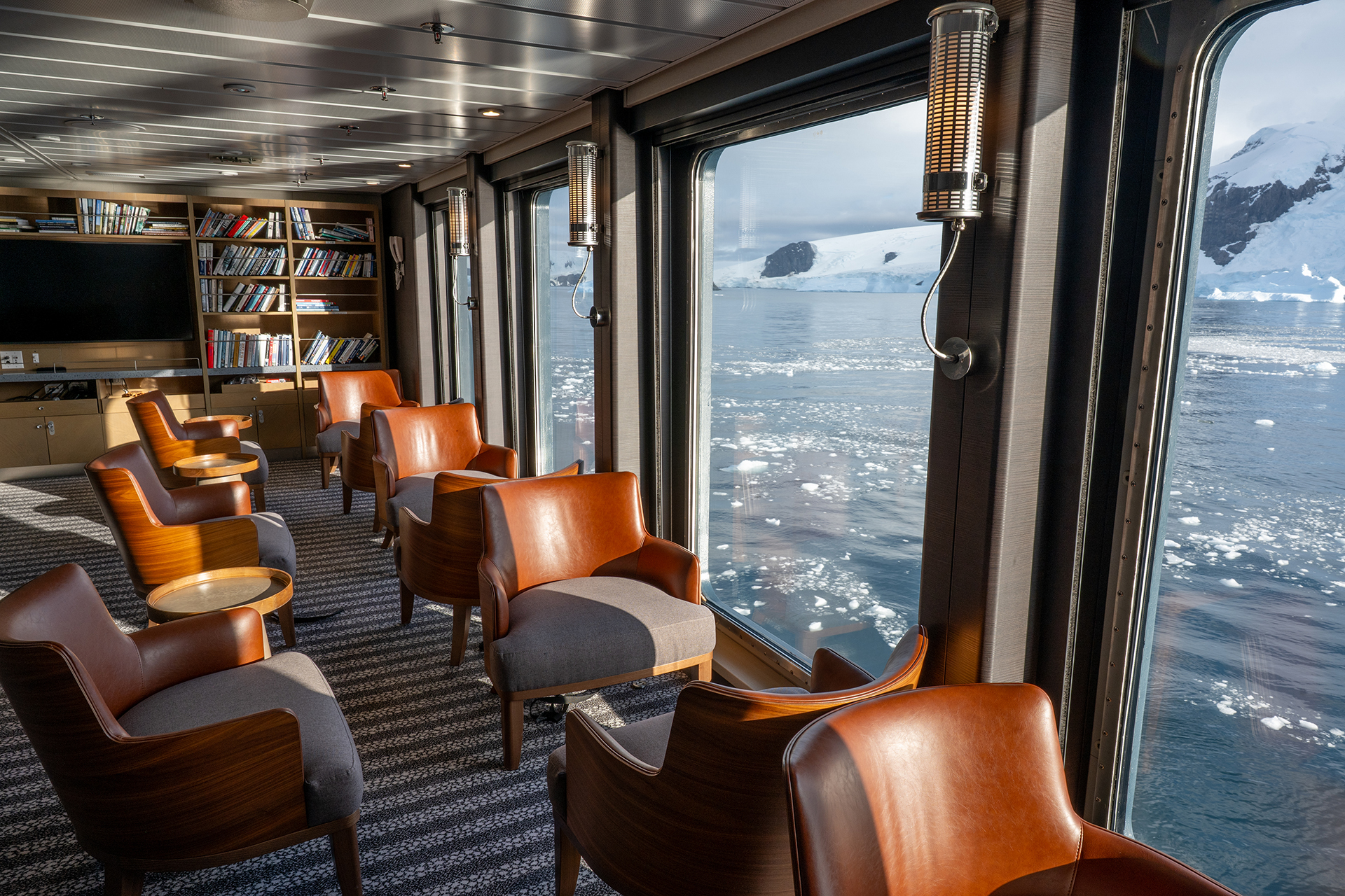library aboard Magellan Explorer polar expedition ship. Leather chairs lines up in front of picture windows showing polar landscape. Books line the shelves on the wall.