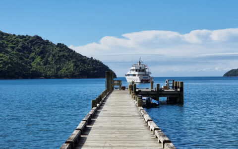 Empty wooden dock leads out to small white yacht Heritage Explorer sitting in calm blue sea by dense green mountains.