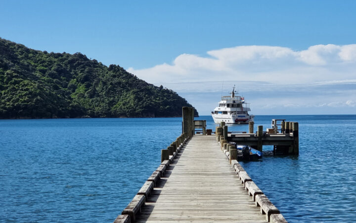 Empty wooden dock leads out to small white yacht Heritage Explorer sitting in calm blue sea by dense green mountains.