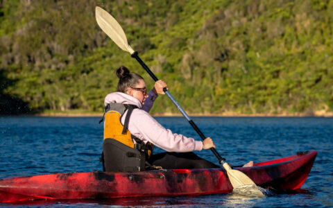 Woman paddles a red kayak in calm blue sea by bright green hills during a Marlborough Sounds cruise.
