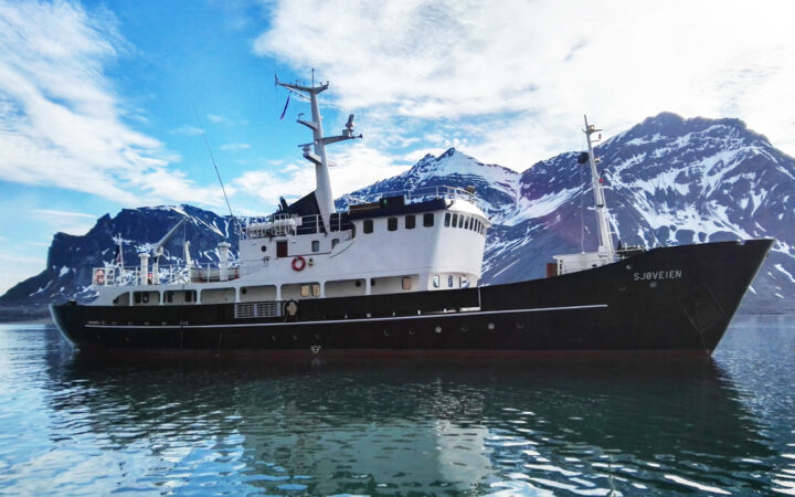 MS Sjoveien Arctic ship with black & red hull, white upper decks & 2 masts, sitting near snowy peaks in calm sea.