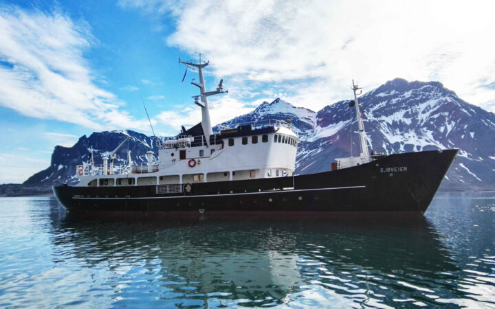 MS Sjoveien Arctic ship with black & red hull, white upper decks & 2 masts, sitting near snowy peaks in calm sea.
