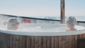 2 women sit in a white hot tub with wooden exterior aboard MS Sjoveien near icebergs on a sunny day in the Arctic.