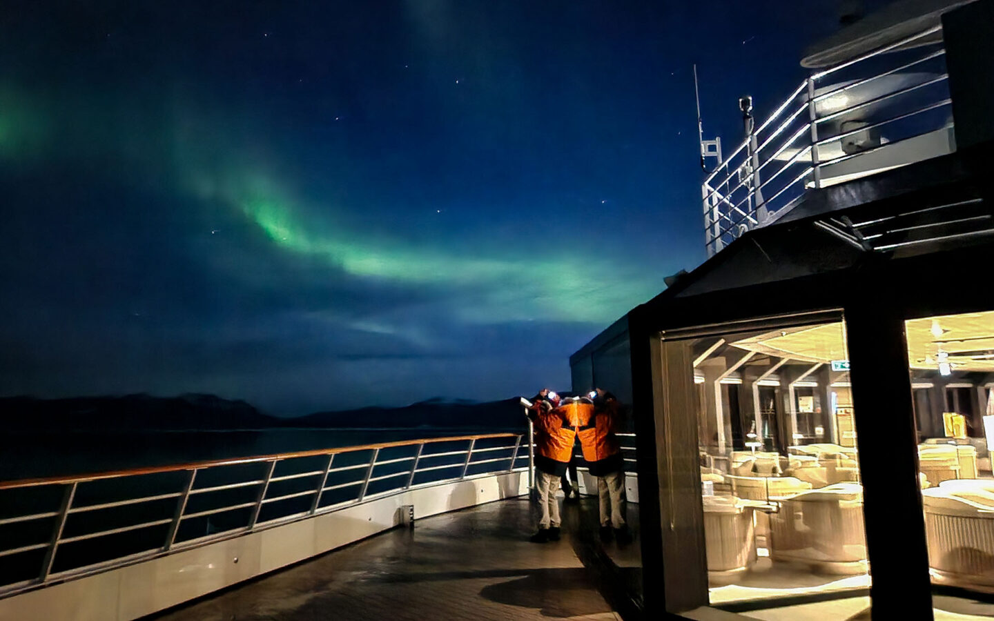 A traveler in an orange jacket stands on the deck of an expedition ship at night taking a picture of the northern lights