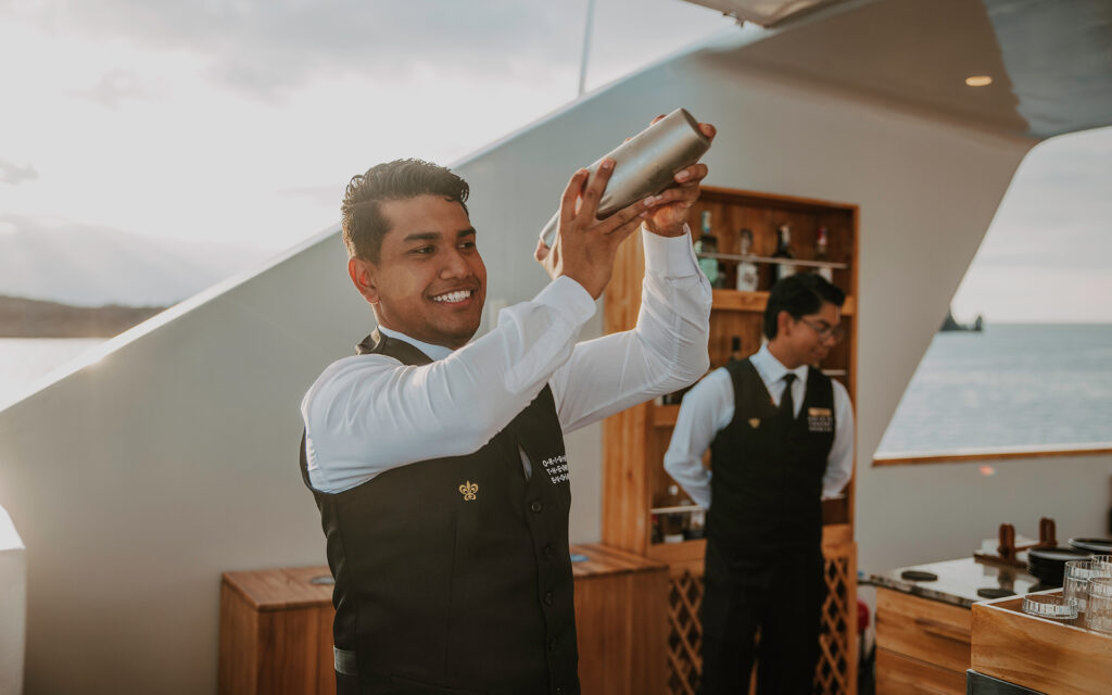 Smiling bartender wearing vest and white collared shirt shakes a cocktail maker Aboard luxury Galapagos cruise Origin and Theory