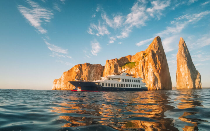 Origin, Theory or Evolve Galapagos luxury yacht with dark blue hull & white upper decks sits in calm seas in front of tan angular rock at sunset.