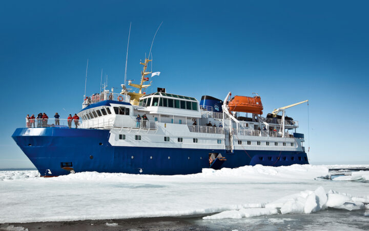 Quest arctic small ship with blue hull, white upper decks & orange life boat sits among ice on a sunny day.