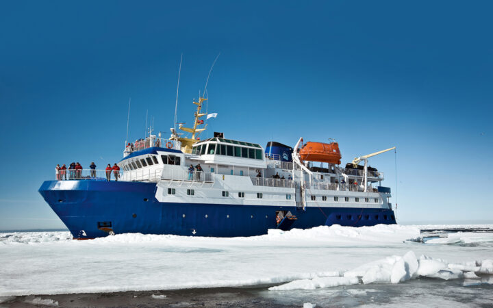 Quest arctic small ship with blue hull, white upper decks & orange life boat sits among ice on a sunny day.