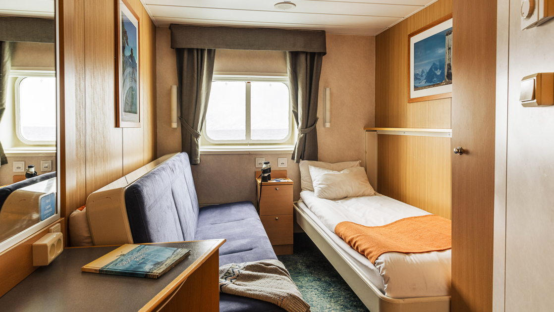 Twin bed in white & salmon-colored linens under reading light, by bedside table & couch in Twin Cabin for Single Use aboard Quest.