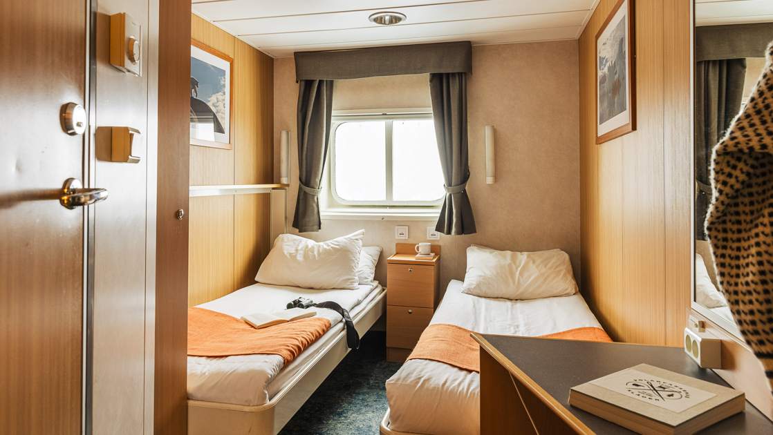 2 twin beds in white & salmon-colored linens by window & wood bedside table in Twin Cabin aboard Quest Arctic expedition ship.