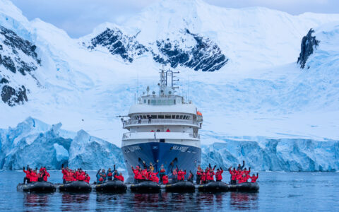 Line of Zodiac boats with Antarctica travelers in red jackets sits in front of Sea Spirit small ship & snowy mountains behind.