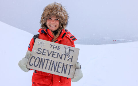 Woman in red parka, white mitts & fake fur hat holds sign reading The Seventh Continent atop a snowfield in an Antarctic storm.