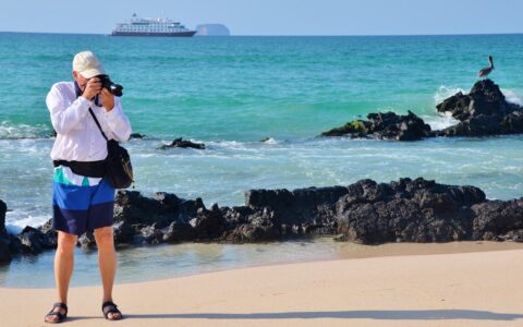 Man stands on a white-sand beach & photographs something out of frame with Santa Cruz II ship & pelican in background.