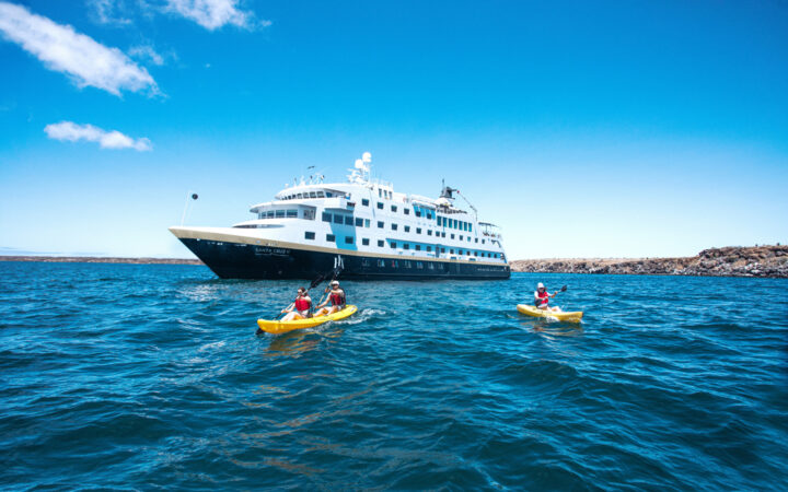 Santa Cruz II Galapagos ship with dark blue hull & gold & white upper decks sits in calm sea as guests paddle kayaks nearby.