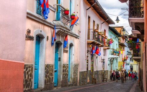 Colorful cobblestone street in Quito, Ecuador, with bright blue doors & multicolored flags flying from small window balconies.