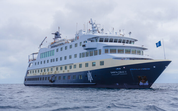 Santa Cruz II Galapagos small ship with dark blue hull & gold & white upper decks cruising on calm waters on a cloudy day.