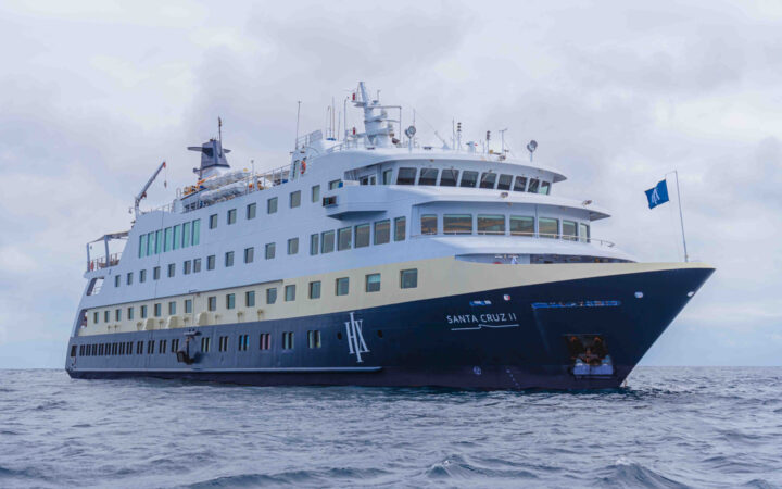 Santa Cruz II Galapagos small ship with dark blue hull & gold & white upper decks cruising on calm waters on a cloudy day.