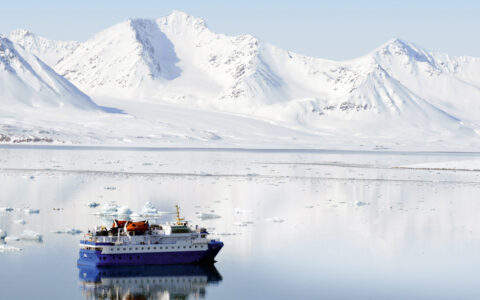 Quest polar expedition ship with blue hull & white upper decks sits in glassy sea by snow-covered mountains in Svalbard.