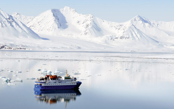 Quest polar expedition ship with blue hull & white upper decks sits in glassy sea by snow-covered mountains in Svalbard.