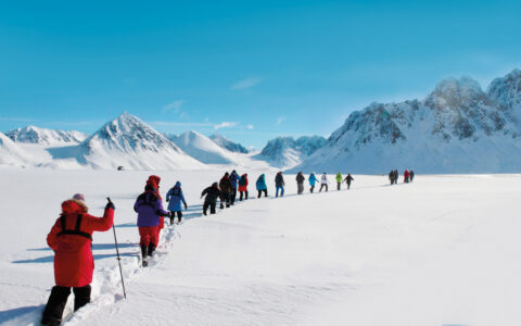 Small group of Svalbard travelers walk a single-file line using snowshoes near snowy peaks on a sunny day in the Arctic.