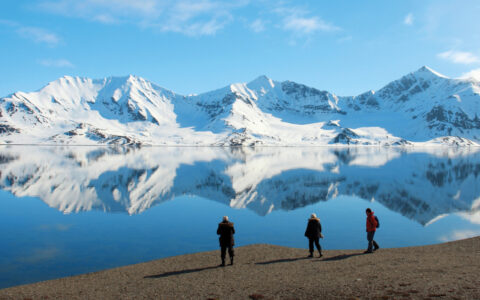 3 Svalbard Adventure Cruise guests walk a rocky overlook above glassy sea & snow-capped mountains in full arctic sunshine.