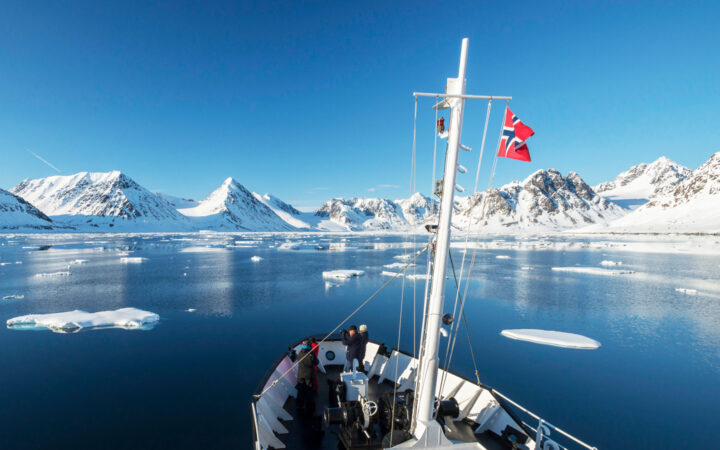A few Svalbard Adventure Cruise guests stand on bow of small white ship, viewing snowy peaks, icebergs & glassy sea in the sun.