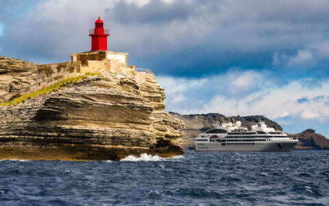 Modern gray & white expedition ship sits by tan cliffs under a red lighthouse during a Mediterranean solar eclipse cruise.