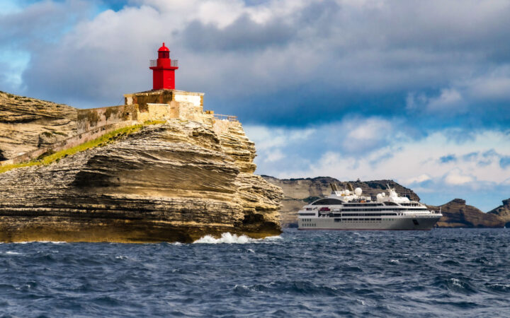 Modern gray & white expedition ship sits by tan cliffs under a red lighthouse during a Mediterranean solar eclipse cruise.