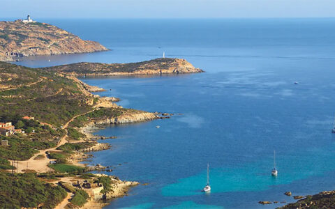 Aerial view of La Revellata, Italy with tan shoreline, white-sand beach & green hills by open ocean with sailboats at anchor.