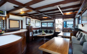 Salon on Katharina sailing ship, with wooden tables, plush seating lining the walls & wooden beams in a patter across the ceiling.