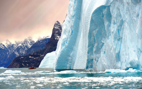 Zodiac cruising by large glacier as sun sets with orange tones against a snowy mountain in the distance on Arctic Wildlife Safari