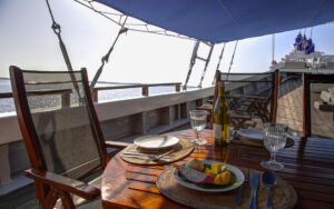 Various sliced fruit, bottle of wine, 2 decorative wine glasses, and a place setting on a table on the deck aboard Ombak Putih yacht