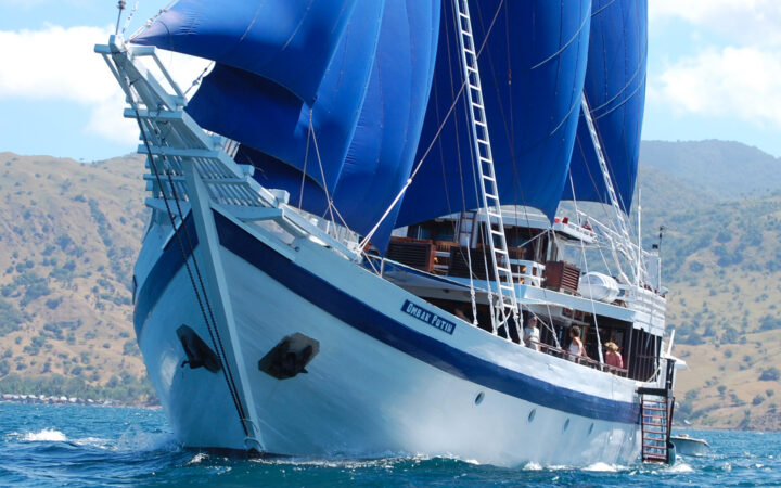 Close up shot of Ombak Putih small ship featuring royal blue sails cruising on a sunny day with land in the background