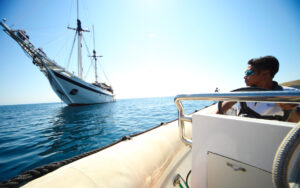 Wide lens photo of Ombak Putih small ship anchored on a sunny day with a guide on a smaller boat in the foreground looking back at it