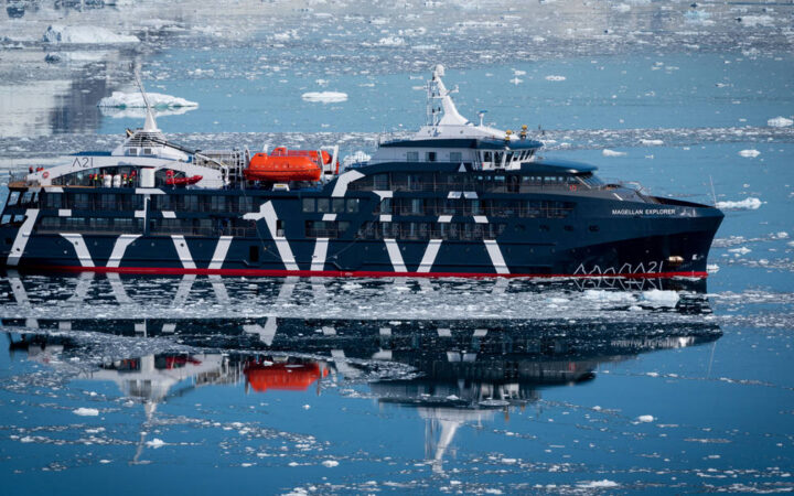Exterior starboard side of dark navy blue Magellan Explorer Antarctica expedition ship floating on icy reflective ocean line.