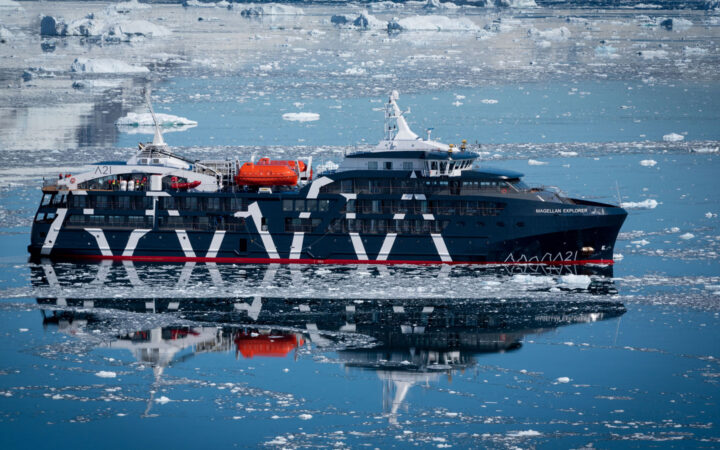 Exterior starboard side of dark navy blue Magellan Explorer Antarctica expedition ship floating on icy reflective ocean line.
