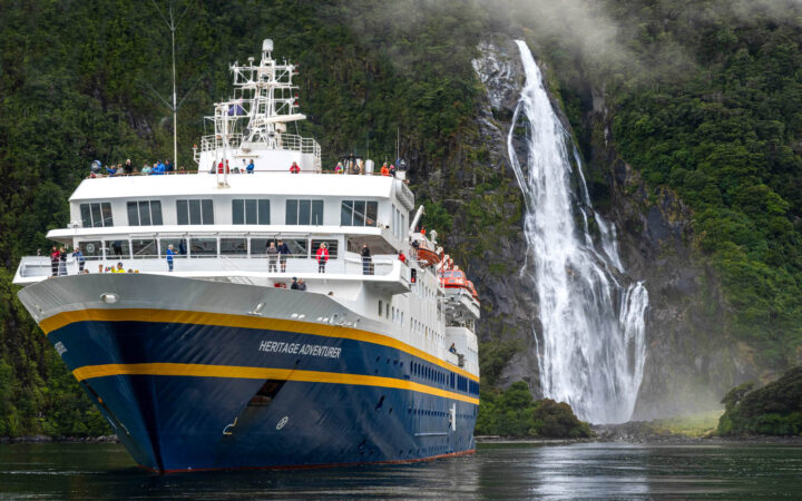 Small ship Heritage Adventurer with blue hull & white upper decks sits beside cascading white waterfall among green cliffs.