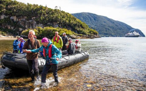 Zodiac boat with Auckland to Queenstown cruise guests in colorful coats comes ashore on a sunny day with a small ship nearby.