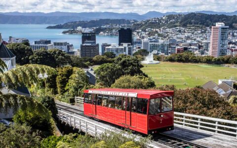 Red trolley car moves up a white track overlooking green grass & downtown with tall urban buildings in Wellington, New Zealand.
