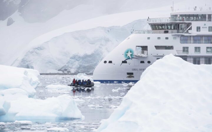 Antarctica travelers ride in a Zodiac through iceberg-filled, calm waters in front of a small white expedition ship.
