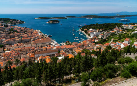 Hilltop view overlooking seaside town in Croatia with green trees, terracotta roofs, teal sea, small harbor & offshore islets.