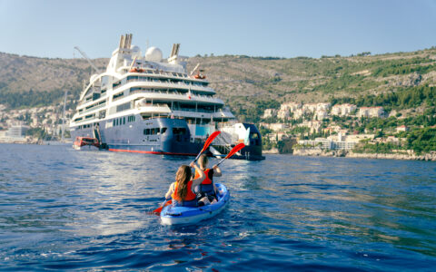 Two people kayaking on calm water in the ocean on a sunny day near a small cruise ship during the Best of Croatia luxury cruise.
