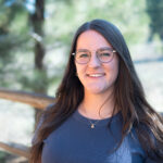 Portrait of Brie Gorter. She's wearing a navy blue shirt and glasses. Pine trees and a wooden fence can be seen in the background.