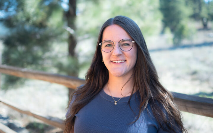 Portrait of Brie Gorter. She's wearing a navy blue shirt and glasses. Pine trees and a wooden fence can be seen in the background.