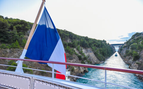 Aft of modern small ship with French flag as it sails the tight Corinth Canal with blue sea & green cliffs on either side.
