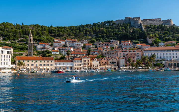 Seaside town in Croatia with terra cotta roofs on beige brick buildings on a green hillside topped by castle walls in the sun.