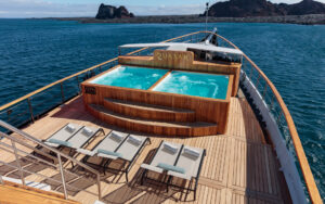 Bubbling Jacuzzis and multiple sunbeds on the deck of small cruise ship, Evolution with oceanic rock formations seen in the distance.